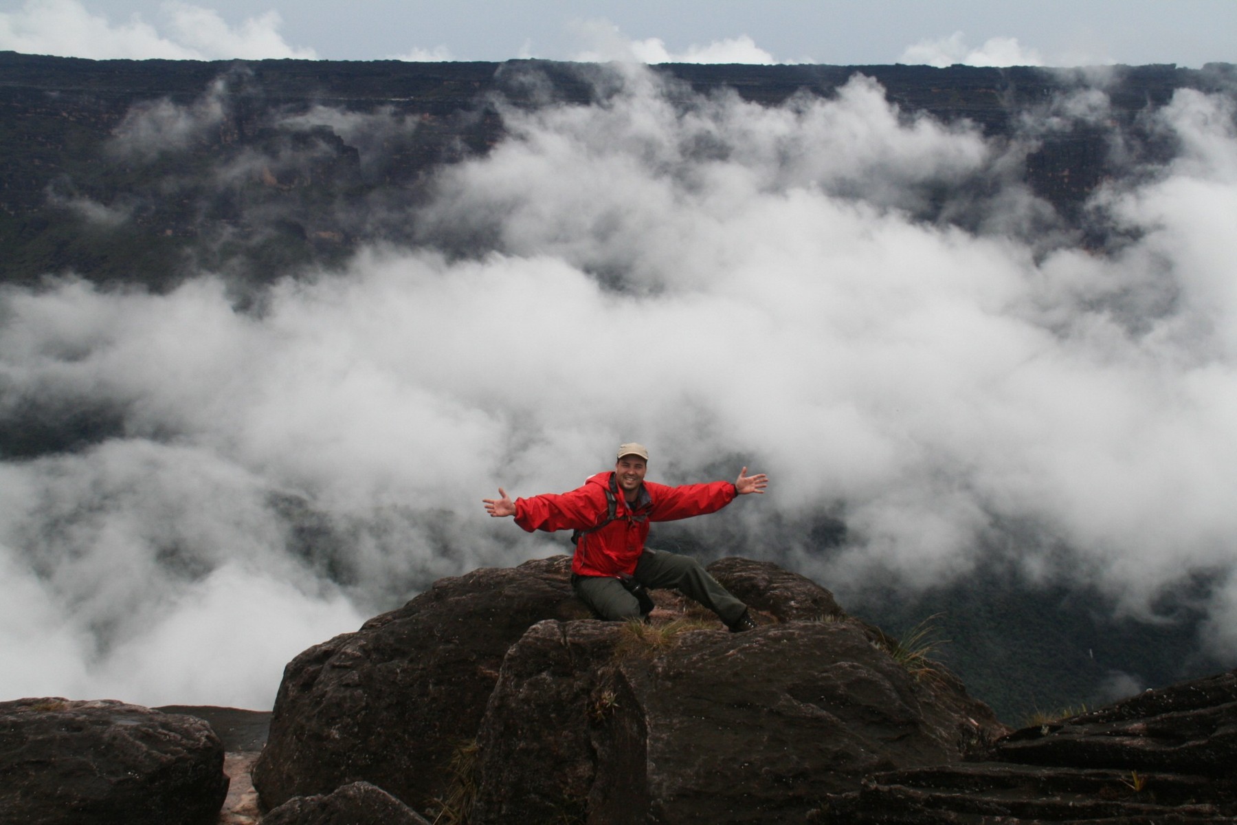 Cristiano de jaqueta vermelha e braços abertos no Monte Roraima, cercado por nuvens.